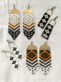 Four pairs of handmade boho beaded fringe earrings made with Japanese seed beads woven onto brass charms. Two pairs are the 'Abiquiu' style in different colors, and the other two are the 'Stella' earrings. Photographed on a beige linen background.