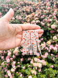 A hand holding a pair of beaded earrings in front of a rose bush