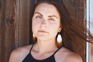 a woman with her brown hair blowing in the wind, wearing an original pair or certain notions handmade beaded fringe earrings in a neutral ombre brown tone