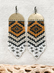The 'Abiquiu' handmade statement fringe beaded earrings by Certain Notions Handmade Designs. Made with Japanese glass seed beads in browns, black and white, woven onto a half-circle raw brass charm. Photographed on a beige linen background.