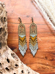 A pair of beaded earrings with browns and creams woven onto a brass cactus charm, photographed on a wooden chest.