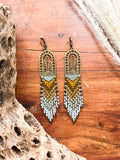 A pair of beaded earrings with browns and creams woven onto a brass cactus charm, photographed on a wooden chest.